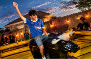 Young man raising one hand while riding a Mechanical Bull at an outdoor evening party.