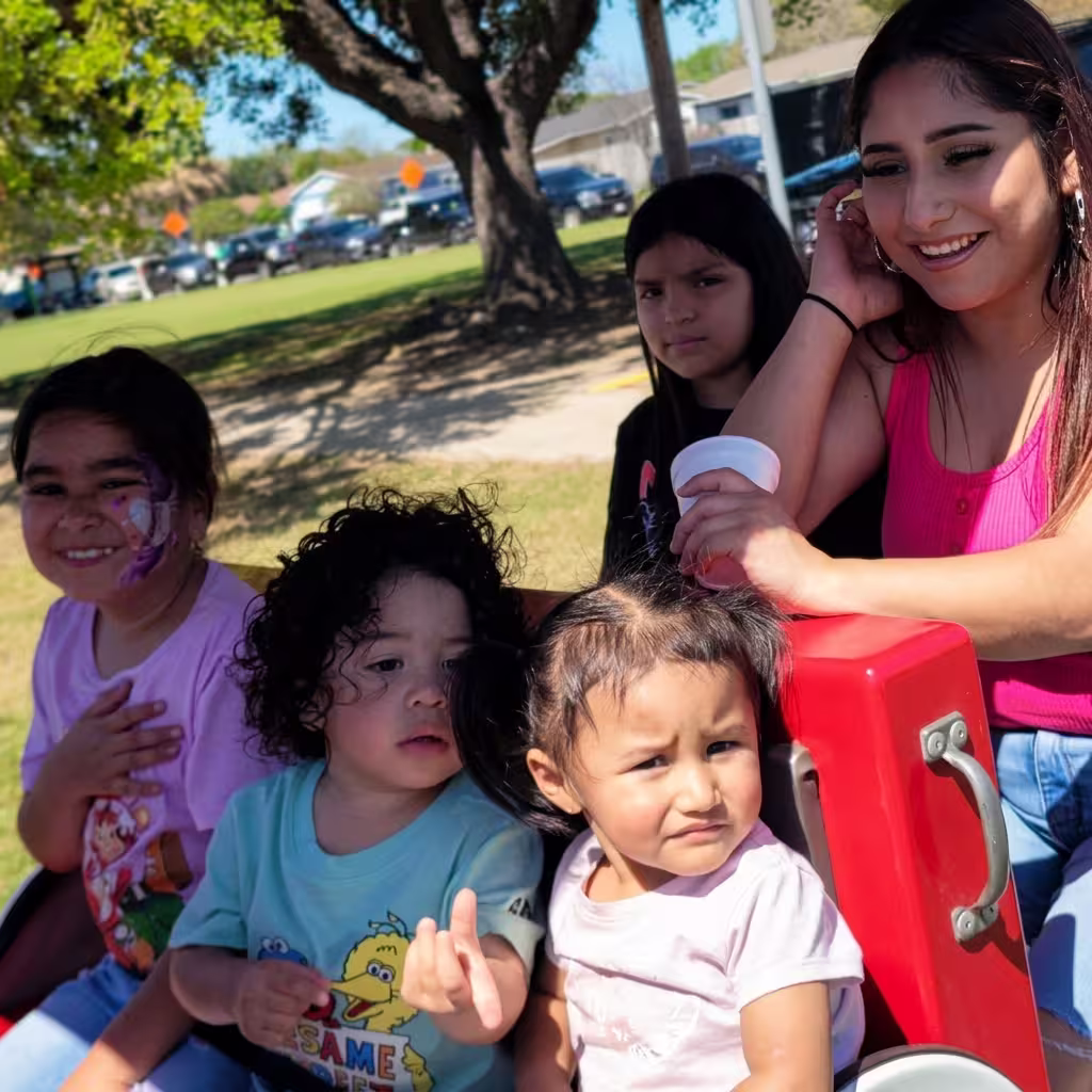 A smiling woman sitting outdoors with four young children, including a girl with face paint and toddlers in a red toy train or ride.